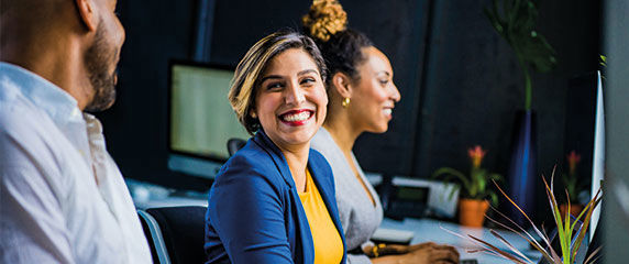 Three office workers sit at a desk. A lady wearing a blue jacket and yellow top is smiling broadly looking at a male co worker to her right. 
